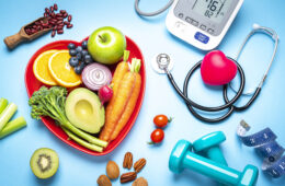 红色心形盘子与新鲜的有机水果和蔬菜拍摄在蓝色背景。盘子旁边有一个数字血压计、听诊器、哑铃和卷尺