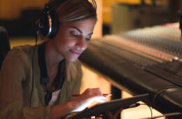照片中，一名女子戴着耳机在工作室的平板电脑上工作，身后是音乐混音板